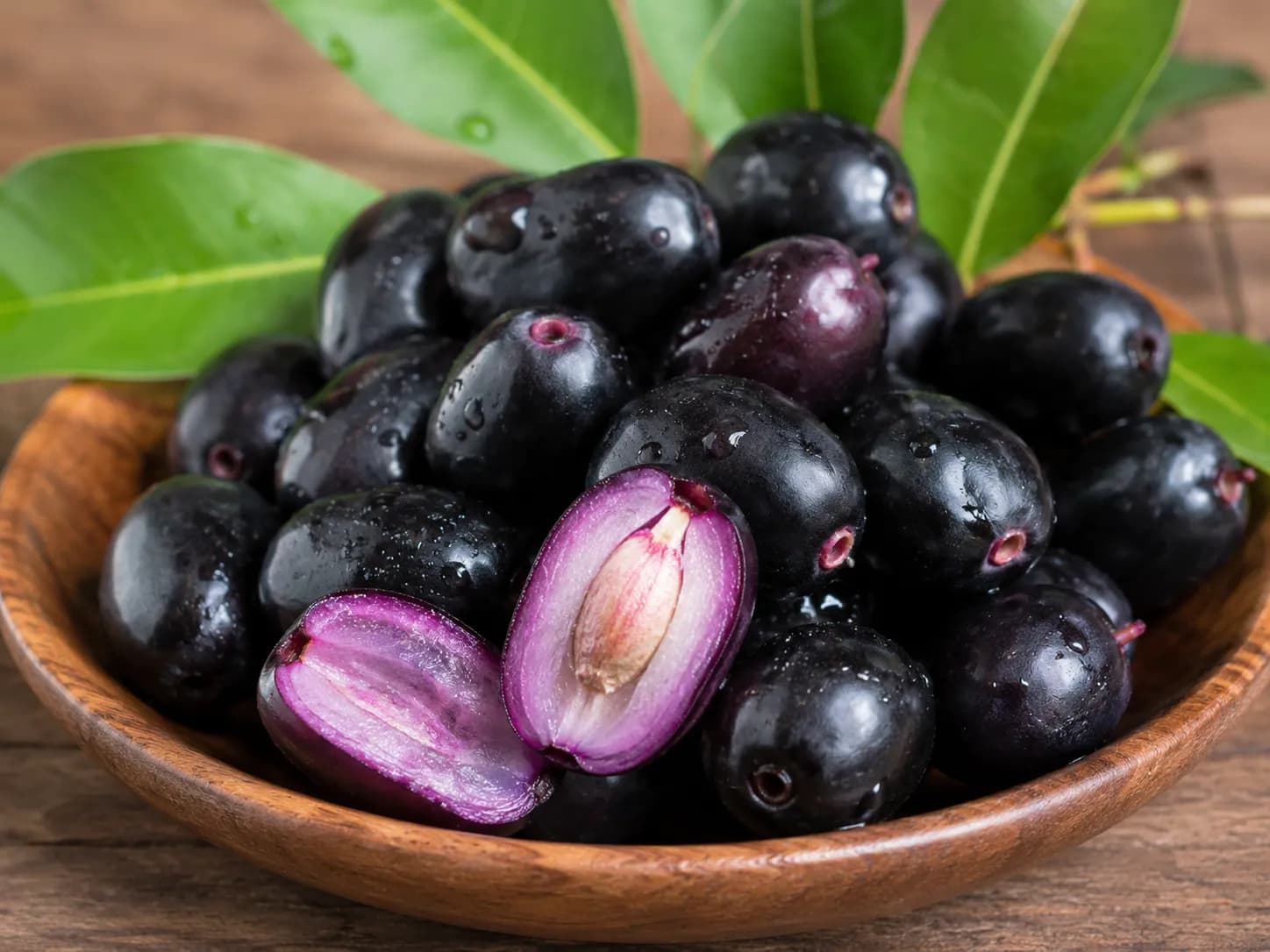 Fresh Black Plum (Jamun) fruits in a wooden bowl with green leaves, featuring a sliced plum showing its purple flesh and seed.